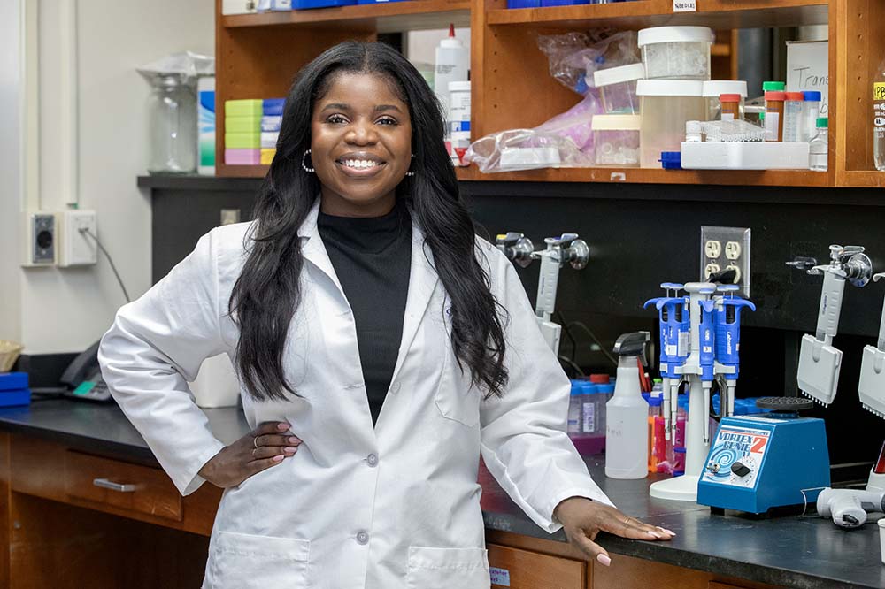 Young woman with long dark hair wearing a white lab coat stands with one hand on her hip and the other on the counter in a laboratory.